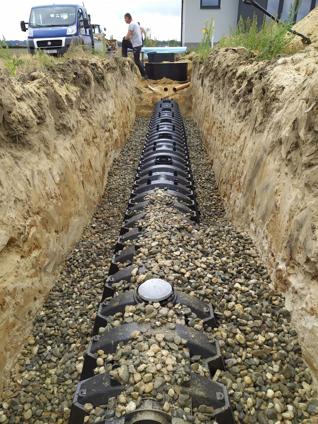 Długi, czarny drenaż rozsączający w wykopie wypełnionym żwirem, widok wzdłuż wykopu, w tle pracownik i zbiornik.