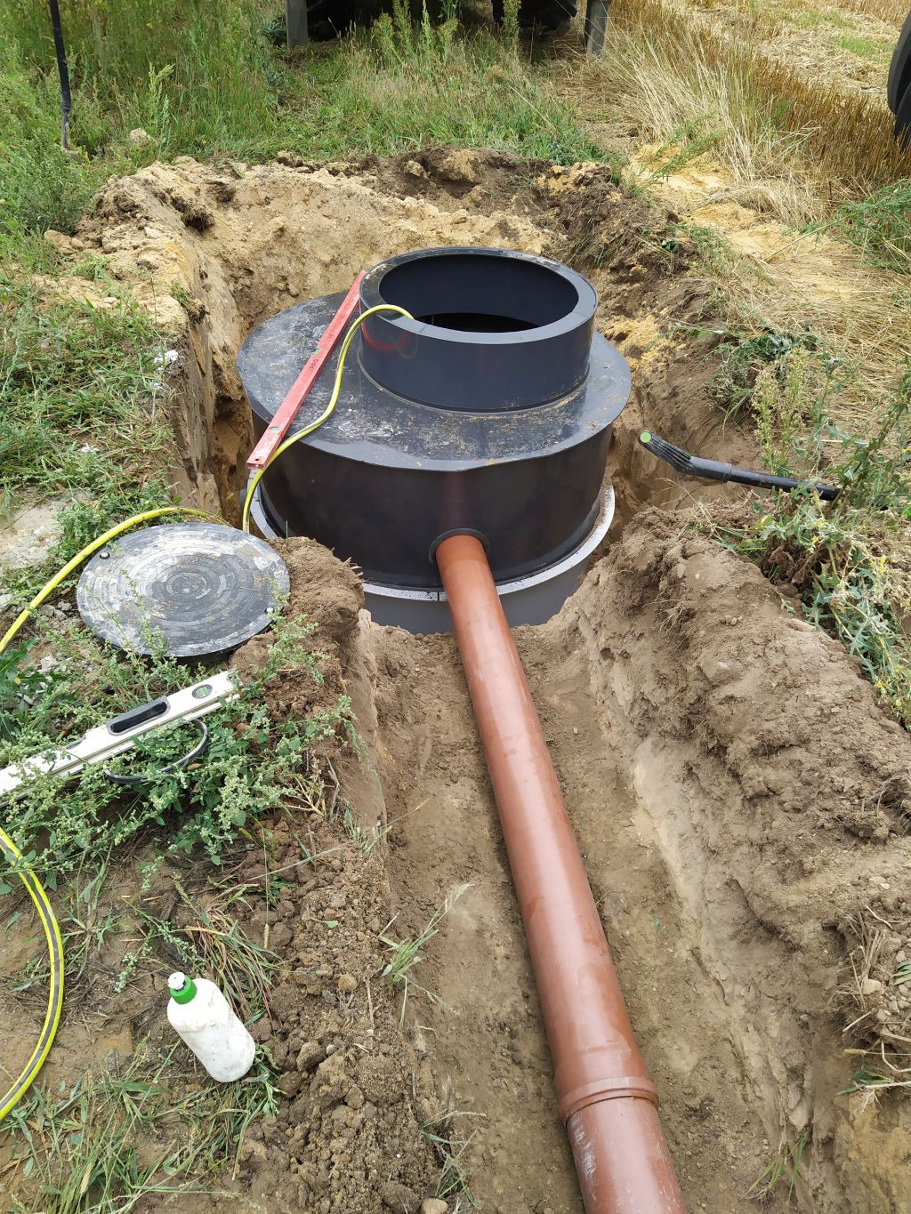 Ciemna, dwukomorowa przydomowa oczyszczalnia ścieków z brązową rurą odpływową, częściowo zakopana w ziemi, z widoczną poziomicą, kablem elektrycznym i butelką kleju obok wykopu.