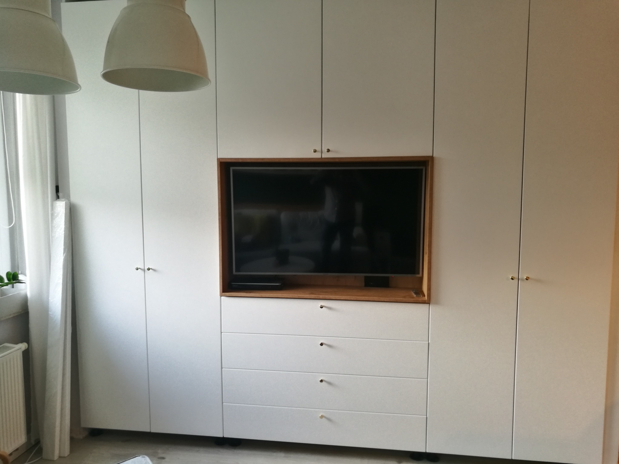Biała szafa z wnęką na telewizor, drewniane obramowanie wnęki, cztery szuflady na dole, minimalistyczne uchwyty, dwie lampy wiszące widoczne w górnym rogu, zasłona w oknie po lewej stronie.