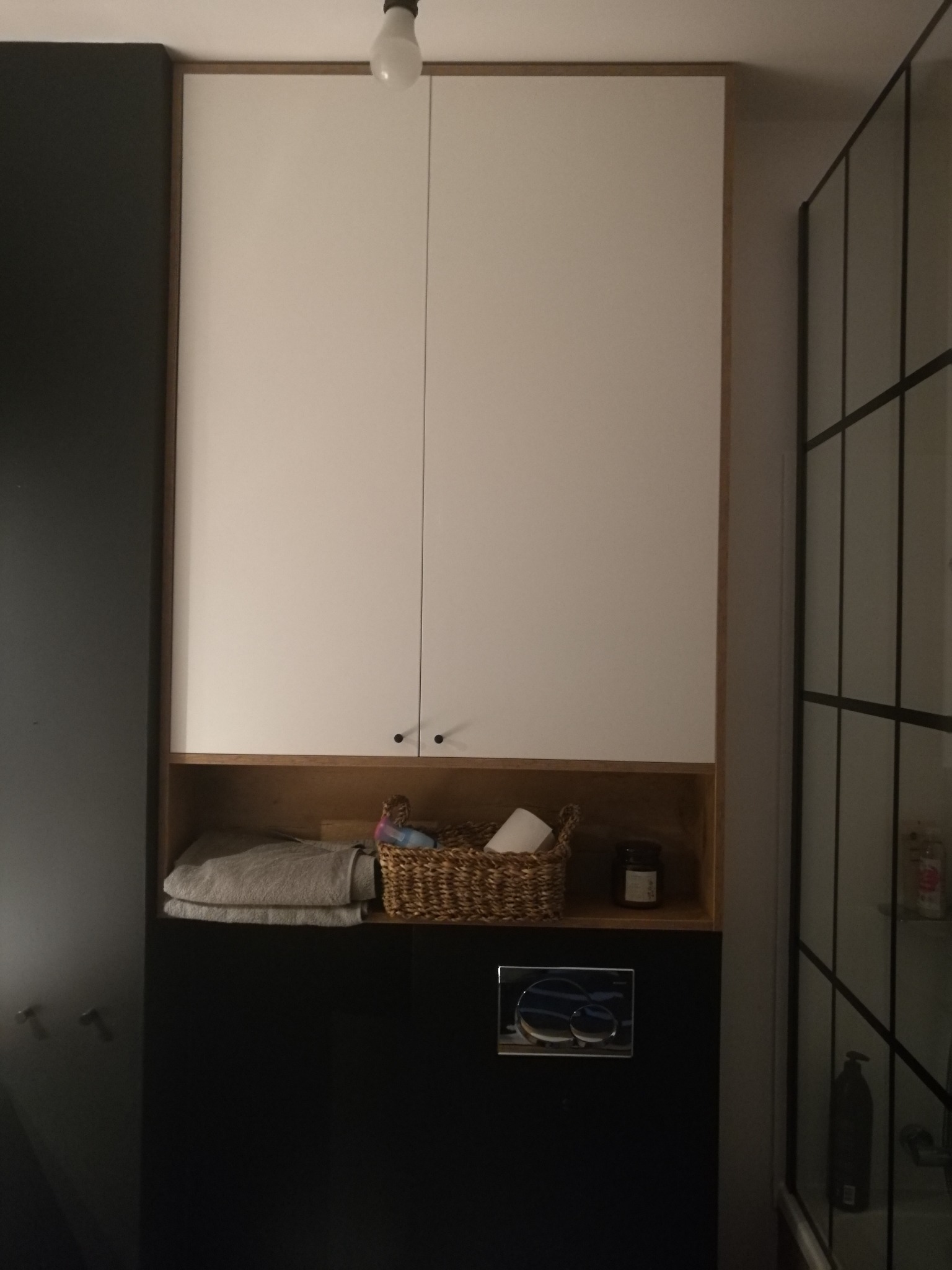 Wysoka zabudowa w łazience z białymi szafkami, otwartą półką z ręcznikami i koszykiem wiklinowym, oraz czarną dolną częścią z przyciskiem spłuczki; kabina prysznicowa z czarnymi szprosami w tle.