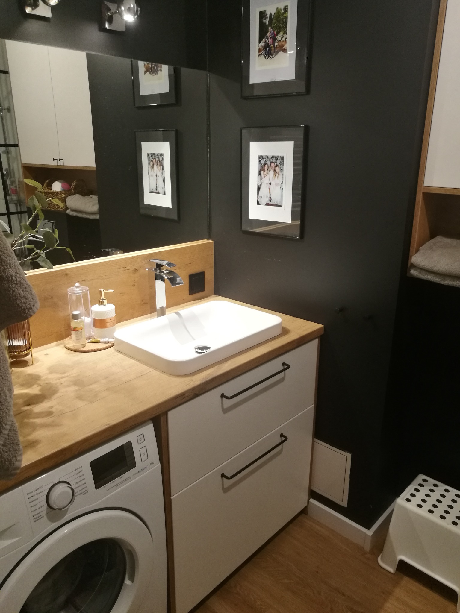 Nowoczesna łazienka z drewnianym blatem, umywalką nablatową, białą szafką z szufladami, pralką pod blatem i czarną ścianą z ramkami zdjęć.