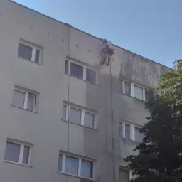 Pracownik na linach zabezpieczających czyści elewację budynku wielorodzinnego za pomocą myjki ciśnieniowej, widoczne różnice w kolorze elewacji w miejscu czyszczenia.