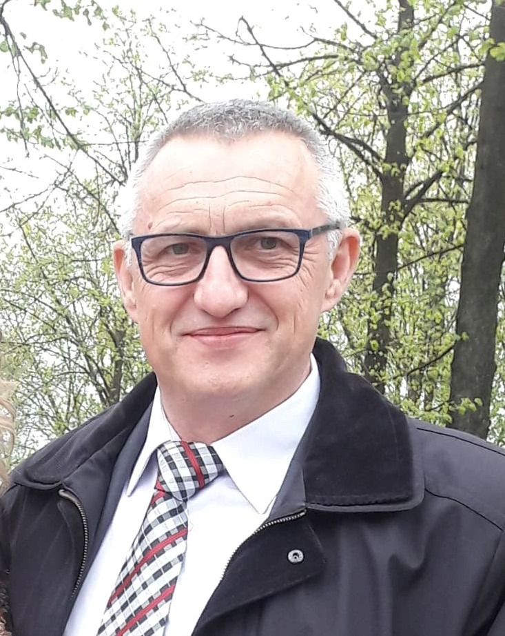 Portret uśmiechniętego mężczyzny w okularach, białej koszuli i krawacie w kratę, ubrany w czarną kurtkę na tle drzew z młodymi liśćmi.