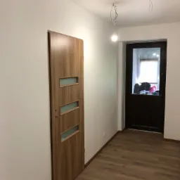 Korytarz po remoncie z drewnopodobną podłogą, białe ściany, wewnętrzne drzwi z przeszkleniami i zewnętrzne drzwi z szybą, widoczne niedokończone podłączenie oświetlenia sufitowego.