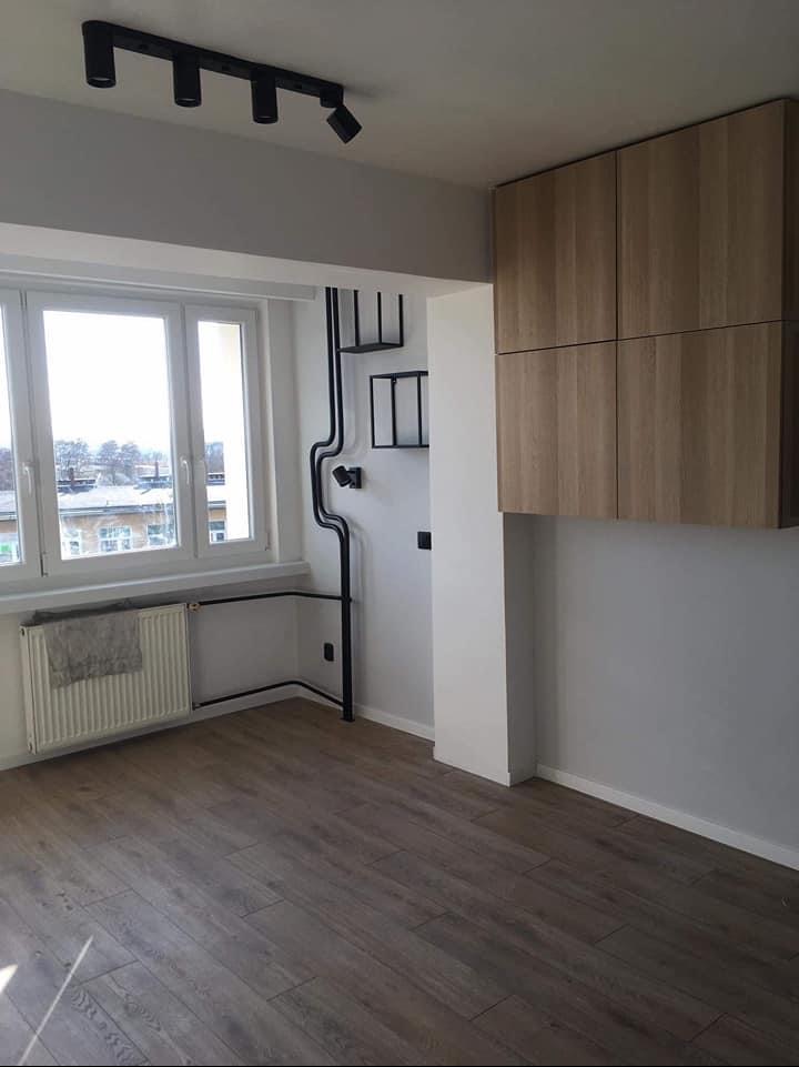 Pokój po remoncie z drewnianą podłogą, szafkami wiszącymi, czarnymi rurami instalacji elektrycznej na ścianie i oświetleniem punktowym.