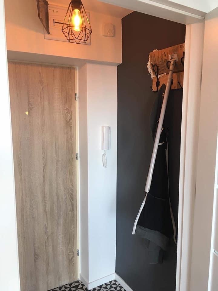 Przedpokój po remoncie: drewnopodobne drzwi wejściowe, czarno-biała mozaika na podłodze, ściana pomalowana na ciemny grafit, wieszak na ubrania i designerska lampa z żarówką Edisona.