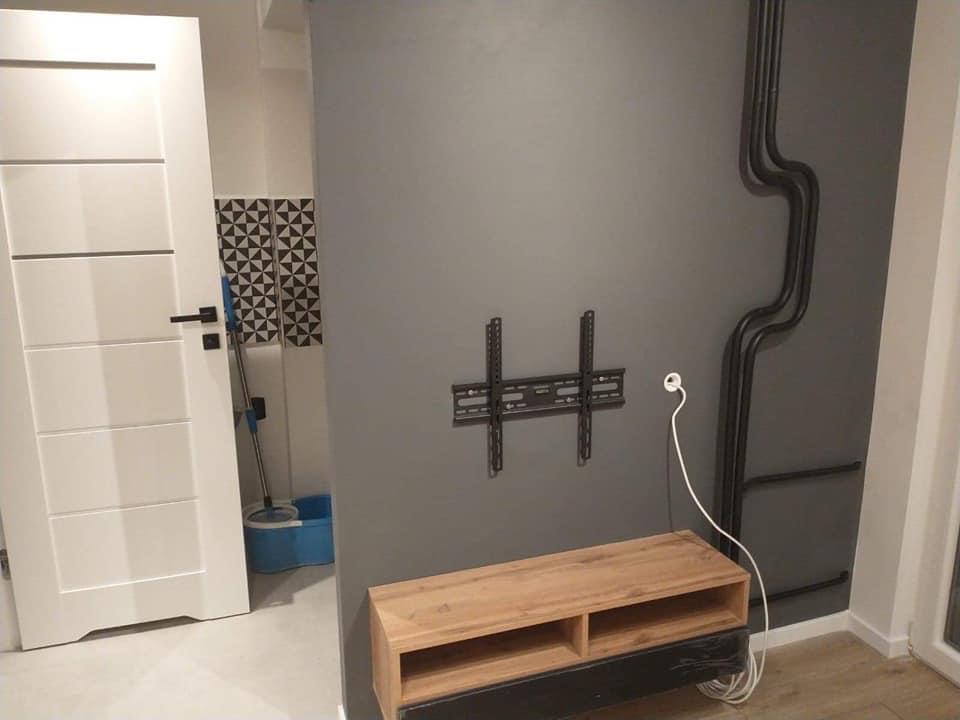 Szara ściana z zamontowanym uchwytem TV, drewnianą szafką RTV i instalacją elektryczną w czarnych rurkach. W tle widoczna biała ściana z mozaiką i mopem.