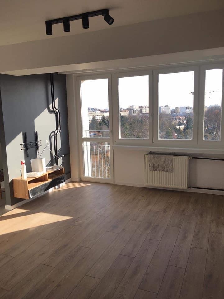 Jasne, przestronne wnętrze z drewnianą podłogą, szarą ścianą z czarnymi rurami, nowoczesną lampą sufitową i widokiem na miasto za oknem.