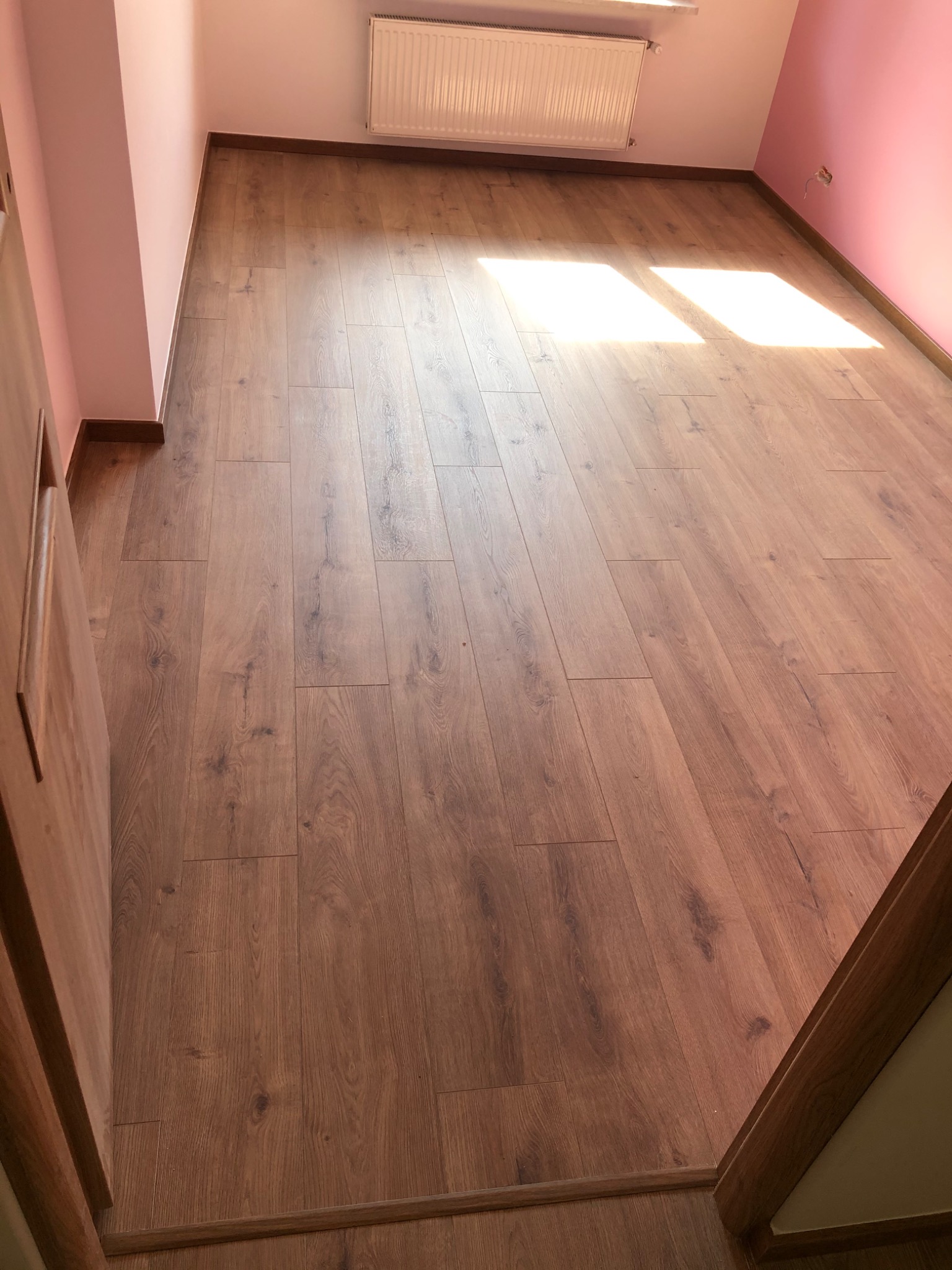 Pusty pokój z panelami podłogowymi w odcieniach brązu, różowe ściany i biały grzejnik pod oknem, wpadające promienie słoneczne.