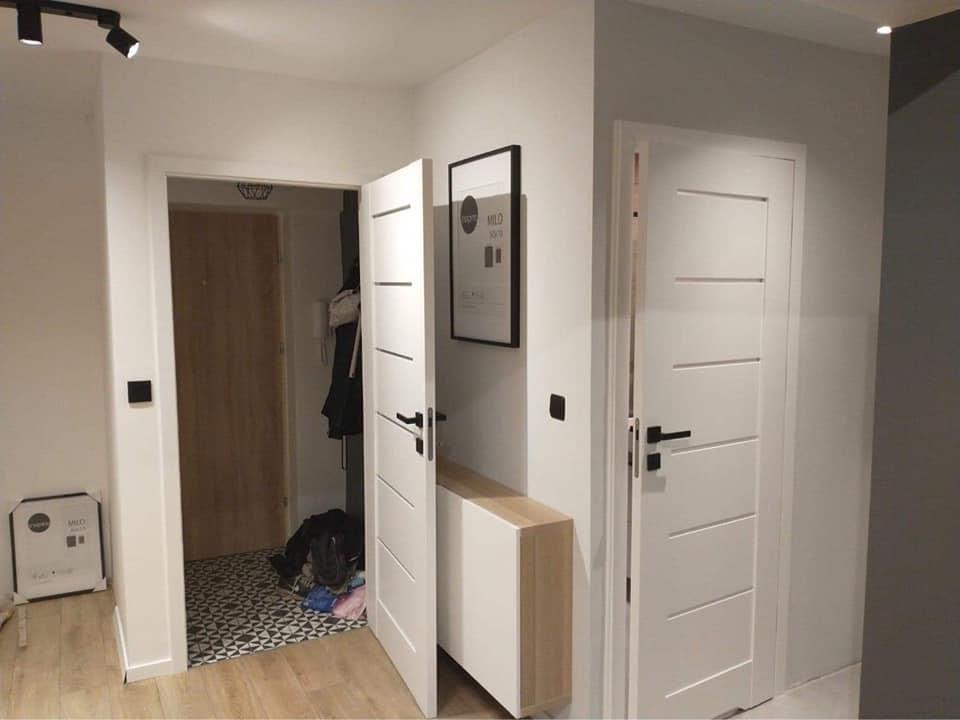 Nowoczesny przedpokój z dwiema białymi, panelowymi drzwiami z czarnymi klamkami, oświetleniem szynowym na suficie i wzorzystą posadzką w głębi pomieszczenia.