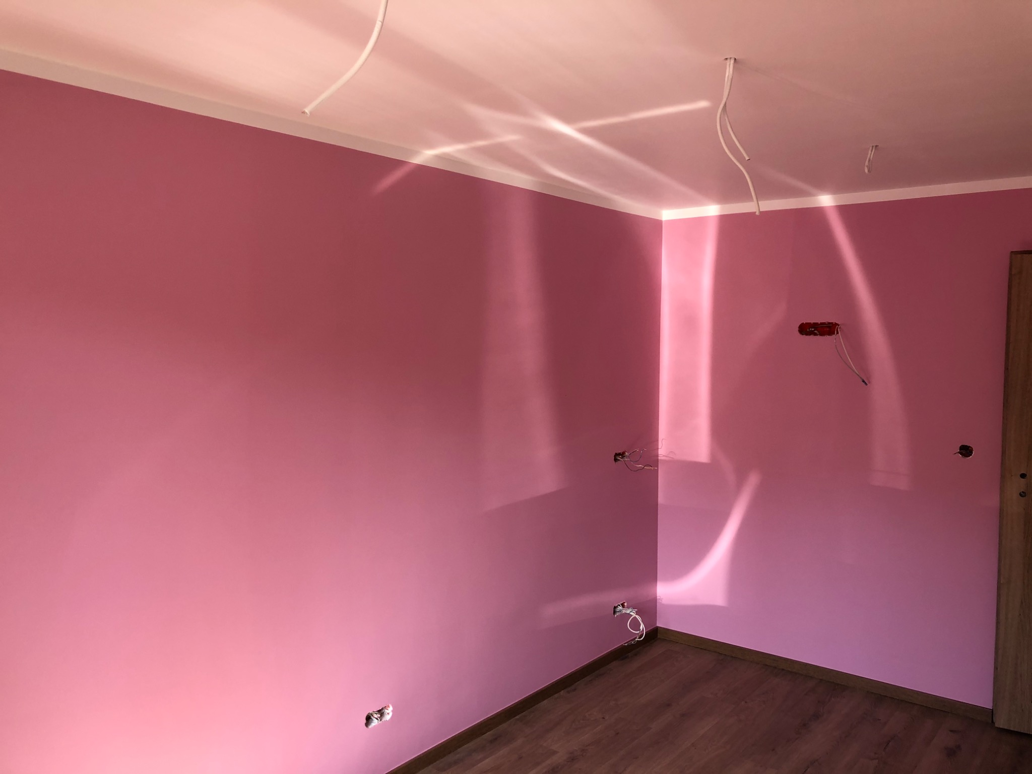 Pokój po malowaniu na różowo, widoczne wystające kable i puszki elektryczne, drewnopodobna podłoga, cień na ścianie.