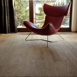 Jasna, drewniana podłoga w Zamościu z wyraźnym usłojeniem, oświetlona naturalnym światłem z okna, na której stoi czerwony fotel na metalowych nogach.
