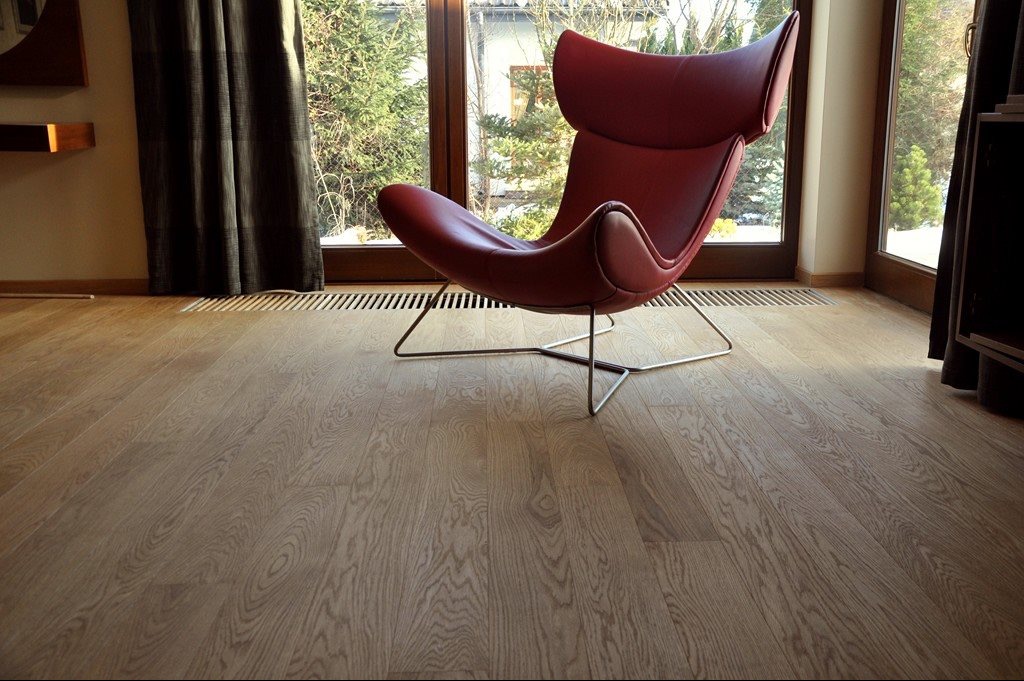 Jasna, drewniana podłoga w Zamościu z wyraźnym usłojeniem, oświetlona naturalnym światłem z okna, na której stoi czerwony fotel na metalowych nogach.