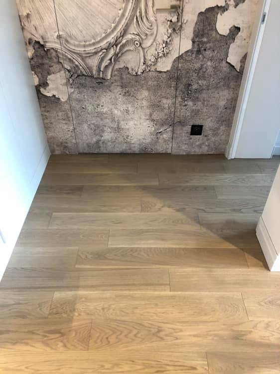 Jasne panele podłogowe ułożone wzdłuż ściany z dekoracyjną tapetą imitującą sztukaterię i spękaną powierzchnię, widoczne gniazdko elektryczne.