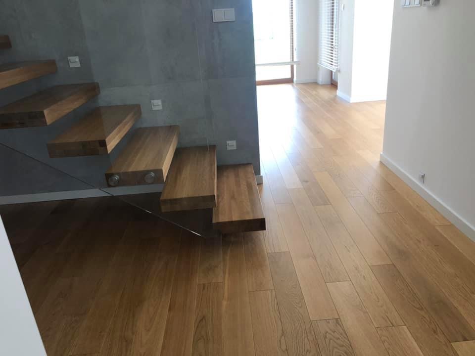 Jasna, drewniana podłoga z parkietu w nowoczesnym wnętrzu, z widocznymi schodami z drewnianymi stopniami i szklaną balustradą, oświetlonymi punktowo.