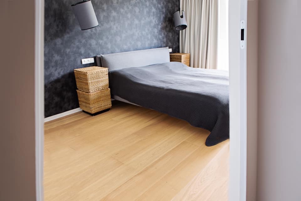 Jasne, drewniane deski podłogowe w sypialni z widoczną częścią łóżka z szarą narzutą, koszykami nocnymi i ciemną ścianą z lampami wiszącymi.