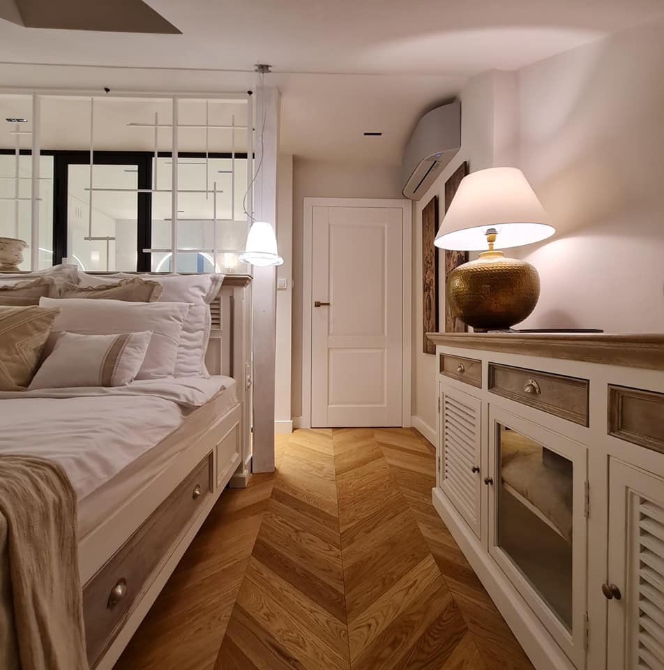 Jasna sypialnia z parkietem ułożonym we wzór jodełki, widok na łóżko z pościelą i szafkę nocną z lampką