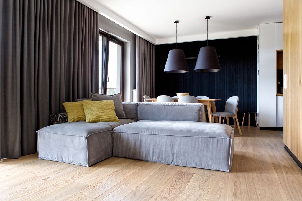 Jasna, drewniana podłoga w nowoczesnym salonie z szarą sofą modułową, żółtymi poduszkami i ciemnymi zasłonami.