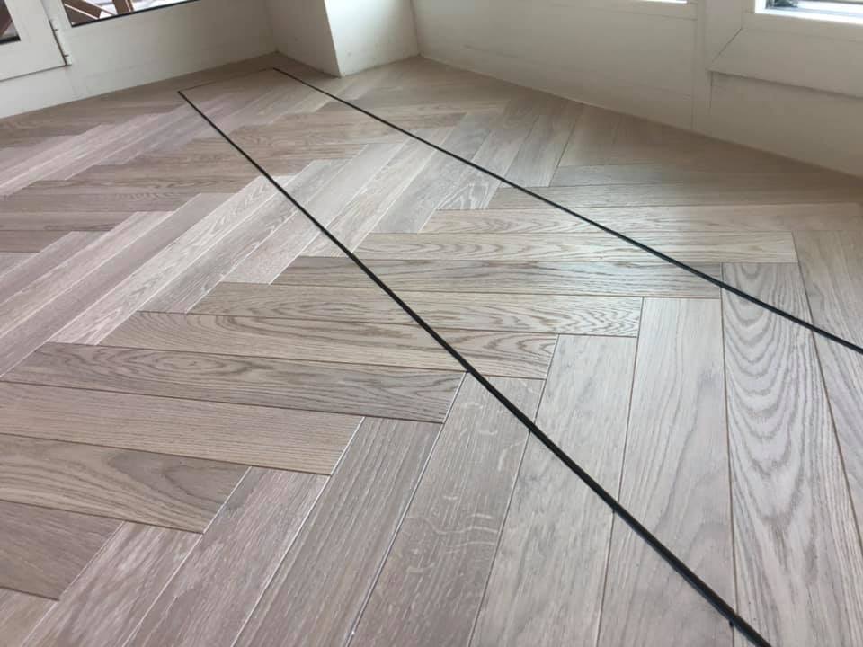 Jasne drewniane deski ułożone w jodełkę, z czarnymi przewodami ułożonymi wzdłuż krawędzi ściany i okna.