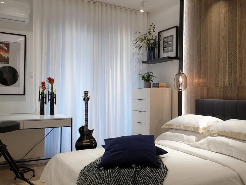 Nowoczesna sypialnia z drewnianą ścianą, białymi zasłonami, czarną gitarą elektryczną stojącą obok biurka i łóżkiem z białą pościelą oraz granatową poduszką.