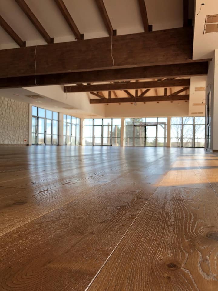 Jasne, przestronne wnętrze z nową, błyszczącą podłogą z drewnianych desek, widoczne promienie słoneczne wpadające przez okna, drewniane belki stropowe.