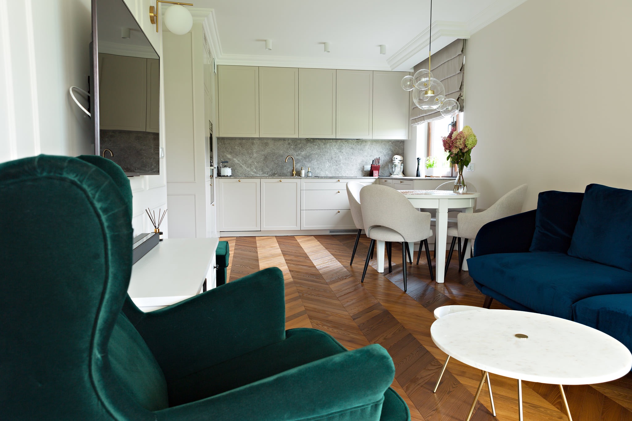 Salon z otwartą kuchnią. Na pierwszym planie fotel w kolorze butelkowej zieleni, parkiet ułożony w jodełkę francuską, w tle zabudowa kuchenna w jasnych kolorach i stół z krzesłami.