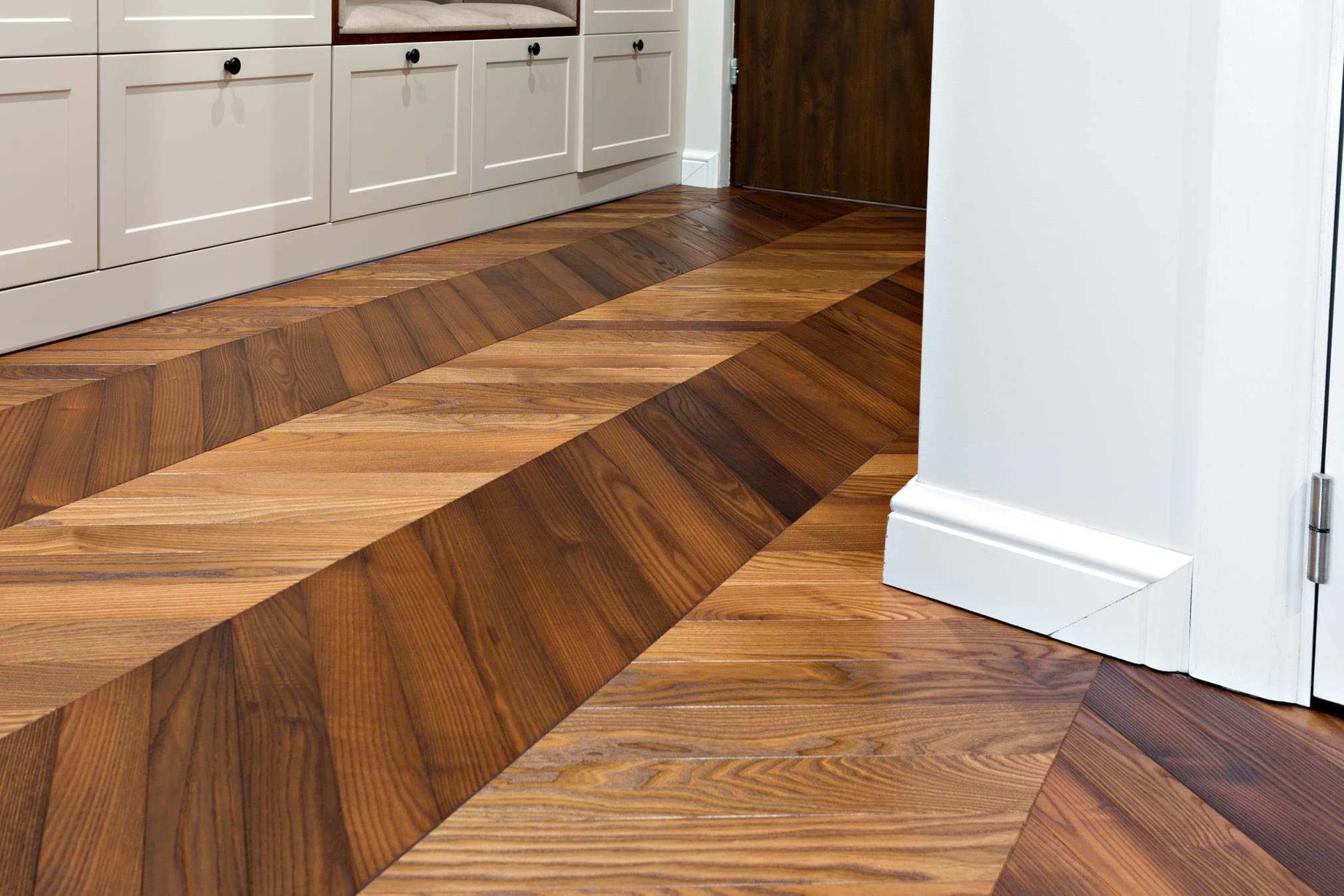 Parkiet ułożony we wzór jodełki francuskiej w odcieniach brązu, sąsiadujący z białą listwą przypodłogową i fragmentem zabudowy kuchennej w kremowym kolorze.