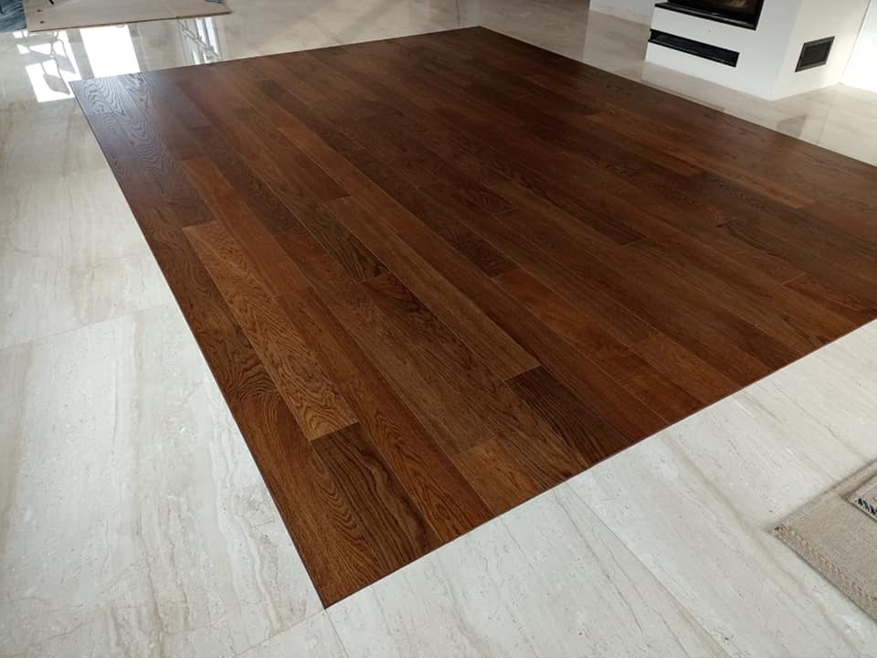 Świeżo położona, ciemna, lakierowana podłoga drewniana z desek, kontrastująca z jasnymi, marmurowymi płytkami podłogowymi w przestronnym salonie z kominkiem.