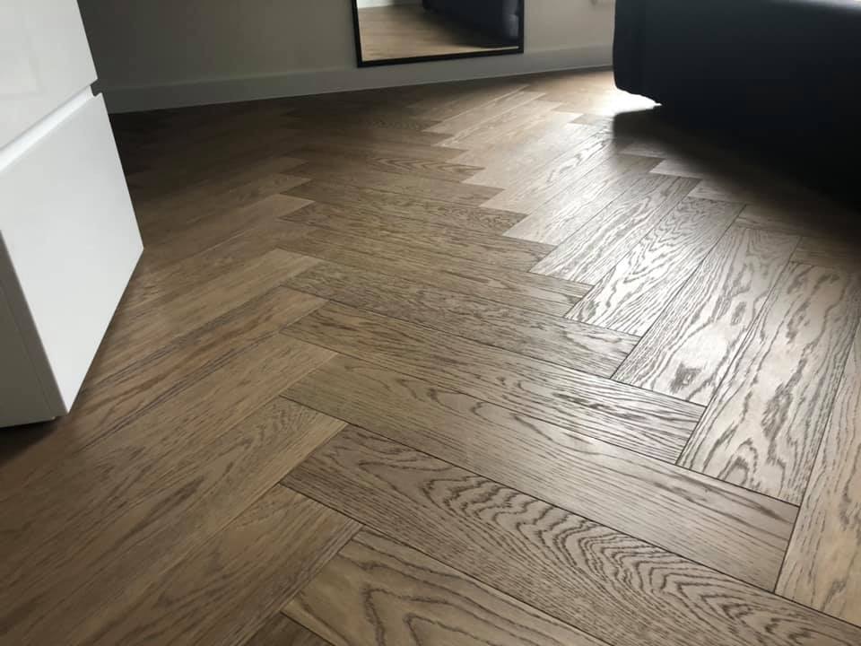 Parkiet ułożony w jodełkę francuską z widocznym usłojeniem drewna, fragment białej szafki i czarnej sofy w tle.