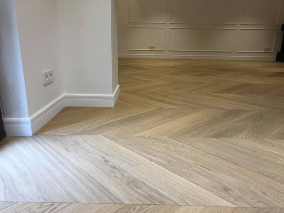 Jasny parkiet ułożony we wzór jodełki francuskiej, białe listwy przypodłogowe, ściana z dekoracyjnymi panelami.