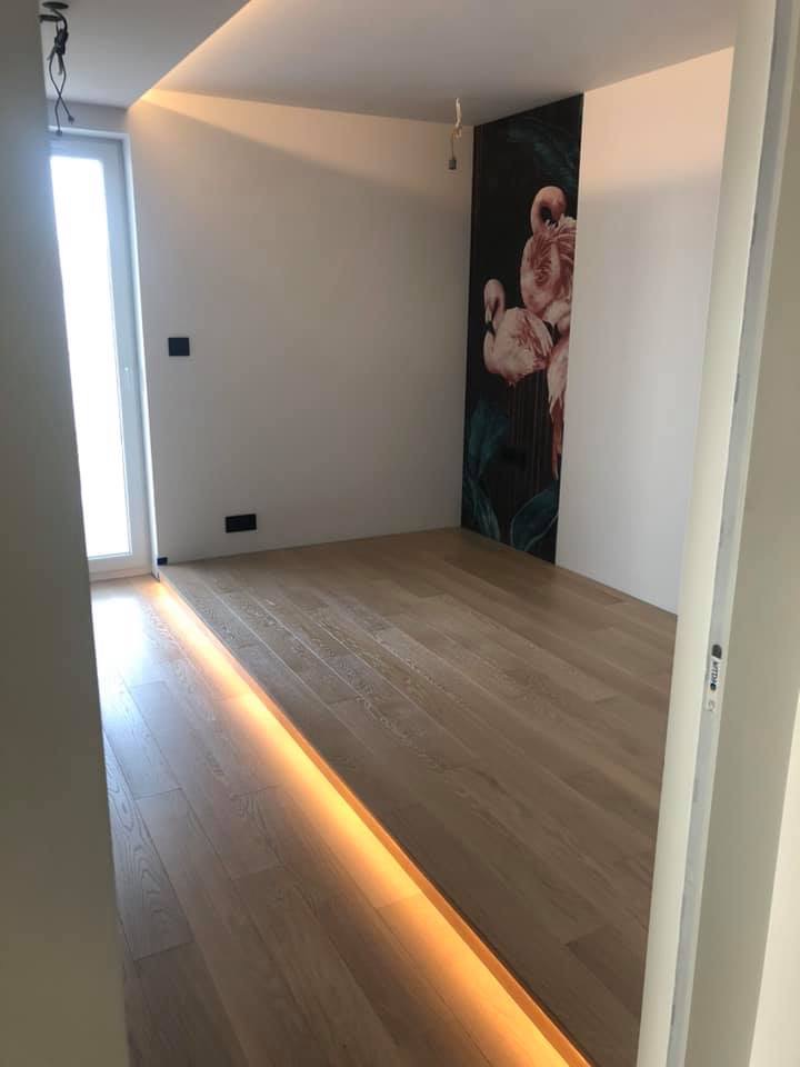 Jasna, drewniana podłoga z listwą przypodłogową podświetloną ciepłym światłem LED w nowoczesnym pokoju z białą ścianą i tapetą z motywem flamingów.