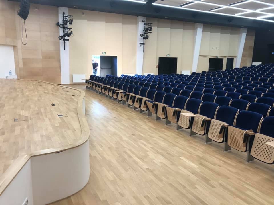 Sala konferencyjna z drewnianą podłogą i rzędami niebieskich krzeseł, oświetlona reflektorami, widok z boku sceny.