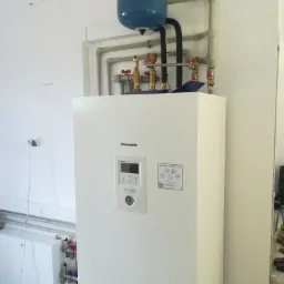 Biała pompa ciepła Panasonic Aquarea z niebieskim zbiornikiem wyrównawczym i instalacją hydrauliczną z zaworami, rurami z tworzywa i metalu, zamontowana w pomieszczeniu z białymi ścianami.