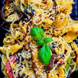 Azjatycka potrawa z makaronem, kurczakiem, warzywami i sezamem, udekorowana świeżą bazylią, podana na czarnym talerzu.