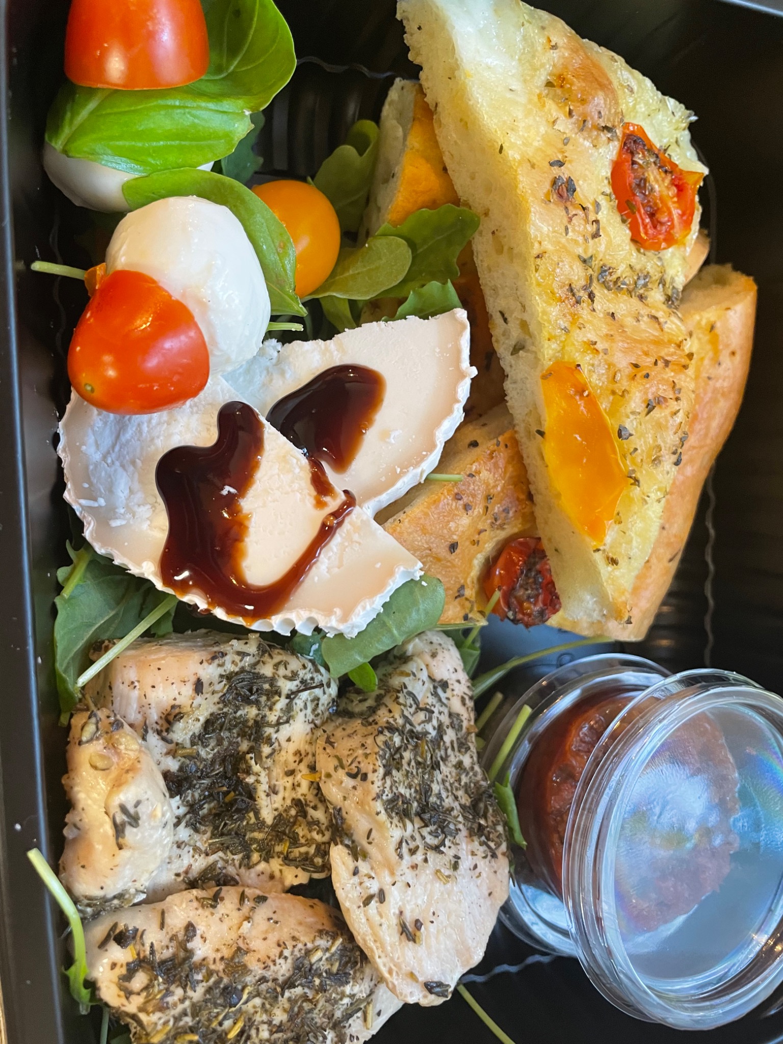 Dietetyczny lunchbox z grillowanym kurczakiem, serem kozim z sosem balsamicznym, mini mozzarellą z pomidorkami cherry i focaccią ziołową, podany na rukoli w czarnym pojemniku.