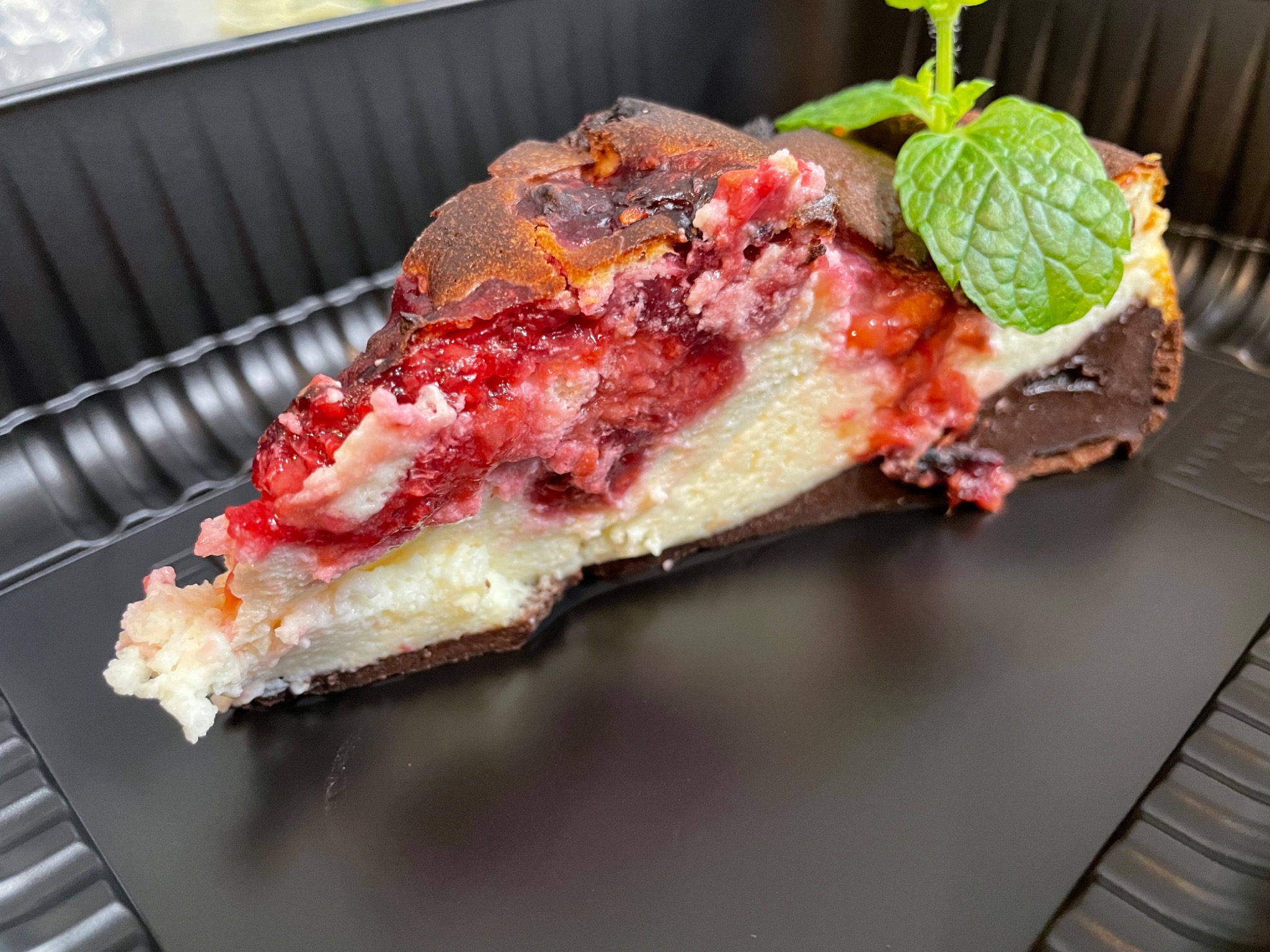 Kawałek sernika baskijskiego z wiśniami, udekorowany miętą, na czarnej, teksturowanej tacy.