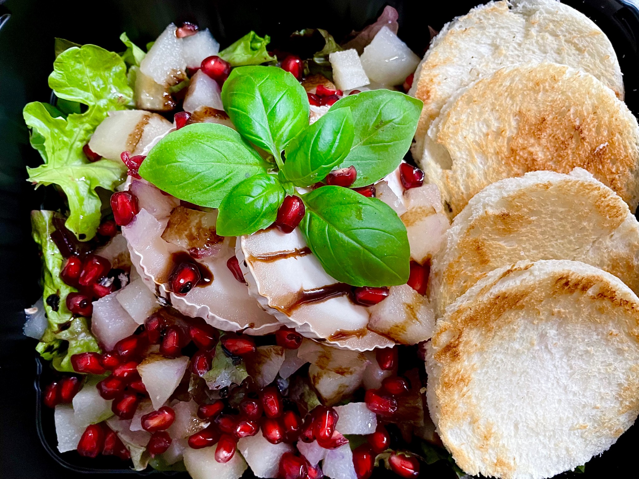 Sałatka z serem kozim, granatem, gruszką i grzankami, polana sosem balsamicznym, udekorowana świeżą bazylią, podana w czarnym pojemniku.