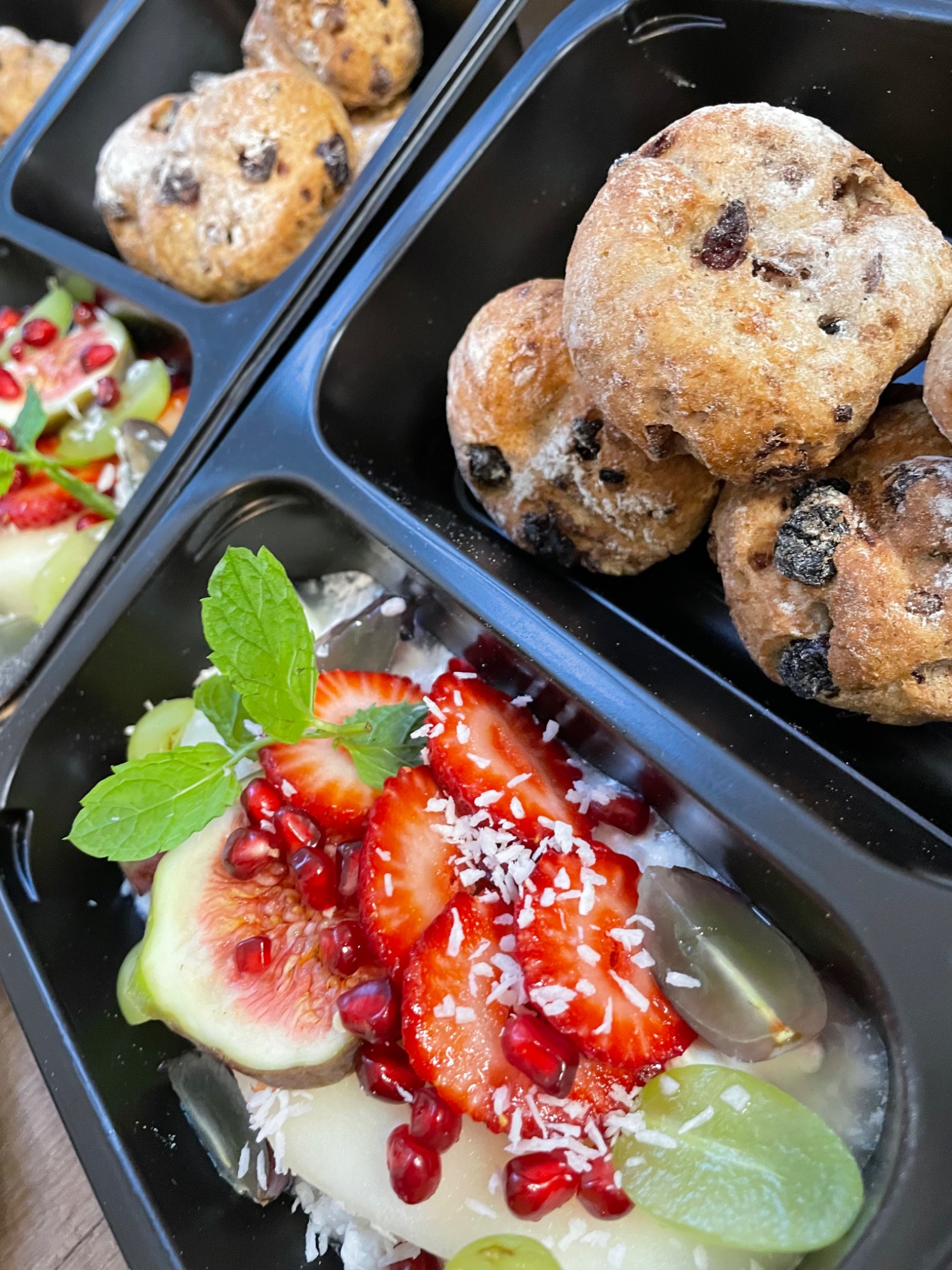 Dwa czarne pojemniki z gotowymi daniami w Krakowie: w jednym bułeczki z rodzynkami posypane mąką, w drugim deser owocowy z figami, truskawkami, granatem, winogronami i miętą, całość posypana...