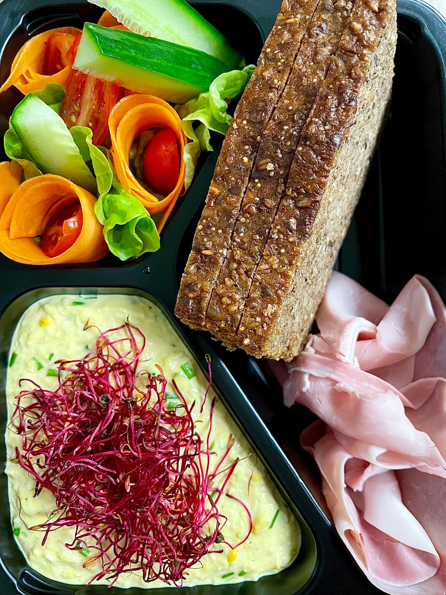 Czarna tacka z przegródkami zawierająca porcję sałatki z rolowanej marchewki i ogórka, kromki ciemnego pieczywa z ziarnami, plasterki szynki oraz pastę jajeczną udekorowaną czerwonymi kiełkami.