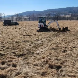 Mini koparka Bobcat w trakcie prac ziemnych na zewnątrz Bolkowa, widoczny samochód w tle, słoneczny dzień.