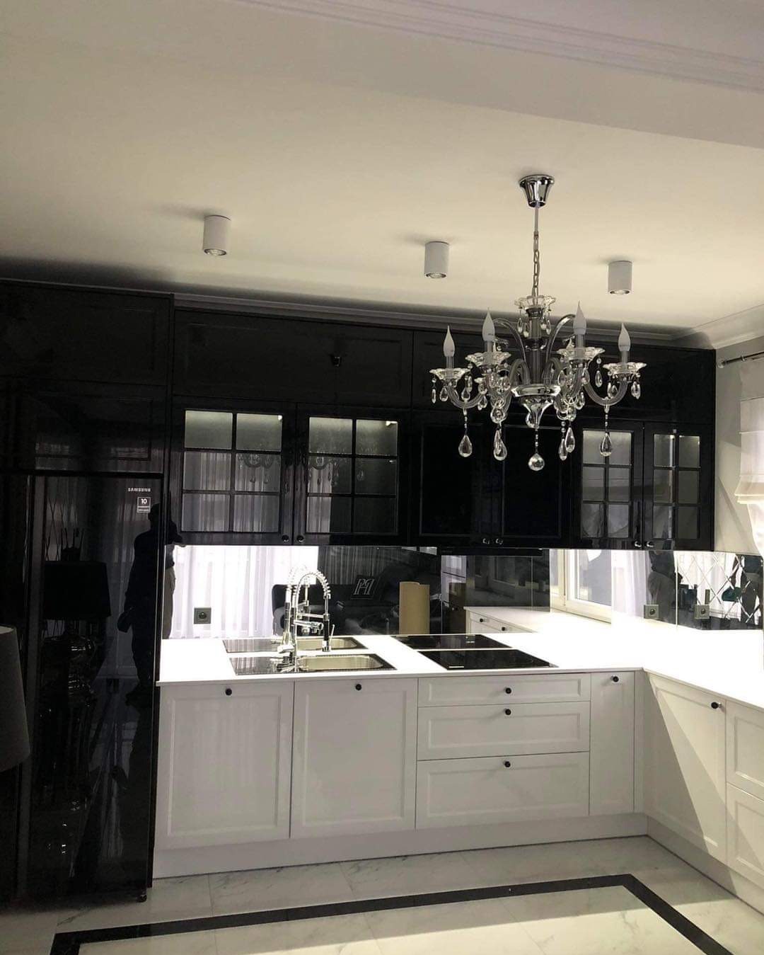 Elegancka kuchnia w czerni i bieli z kryształowym żyrandolem, fronty szafek lakierowane na wysoki połysk, marmurowa podłoga z czarnymi akcentami, zabudowa kuchenna pod sufit.