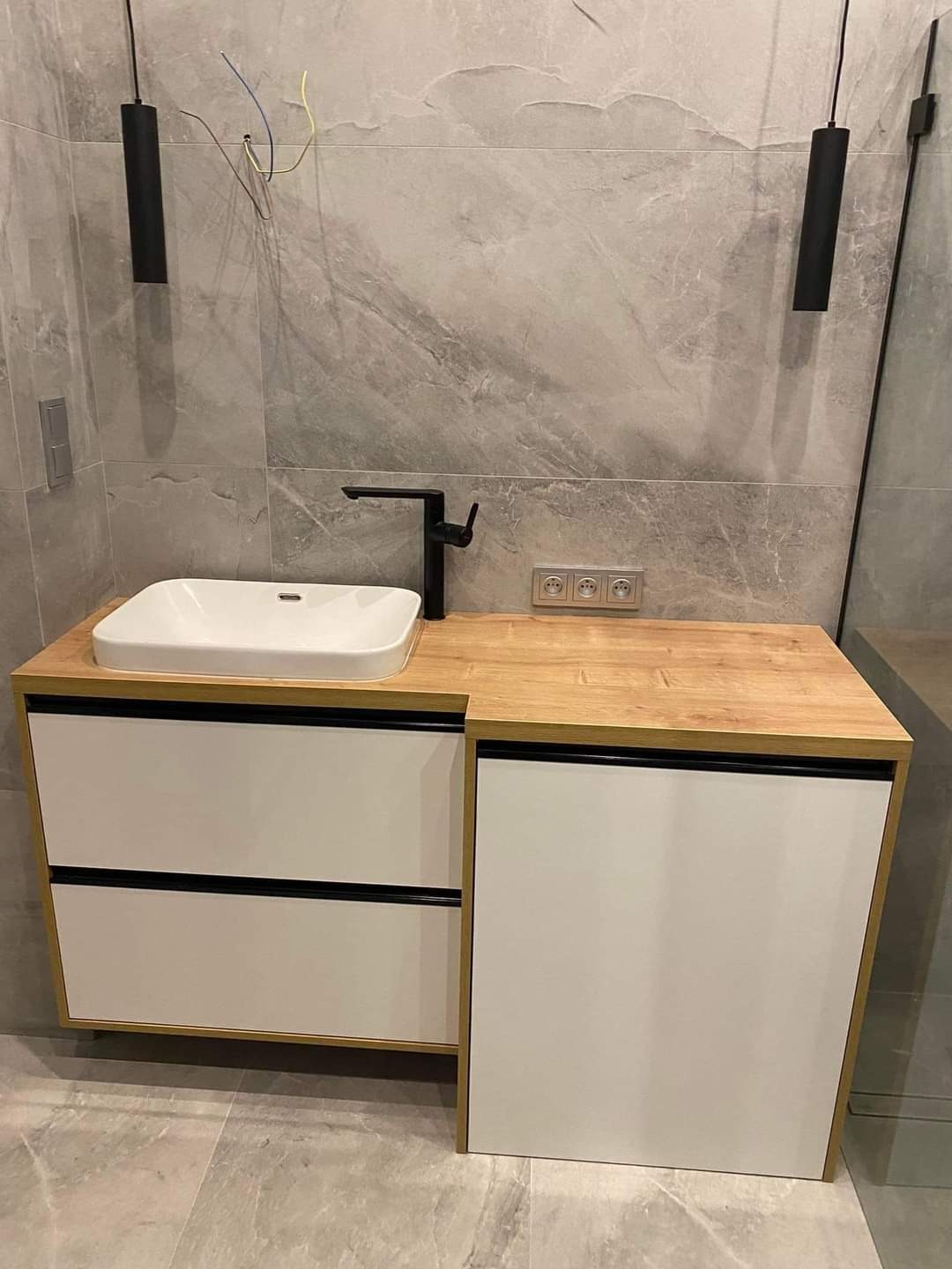 Nowoczesna szafka łazienkowa z białymi frontami i drewnianym blatem, czarna armatura i lampy wiszące, szare płytki imitujące marmur, widoczne niedokończone podłączenie elektryczne.