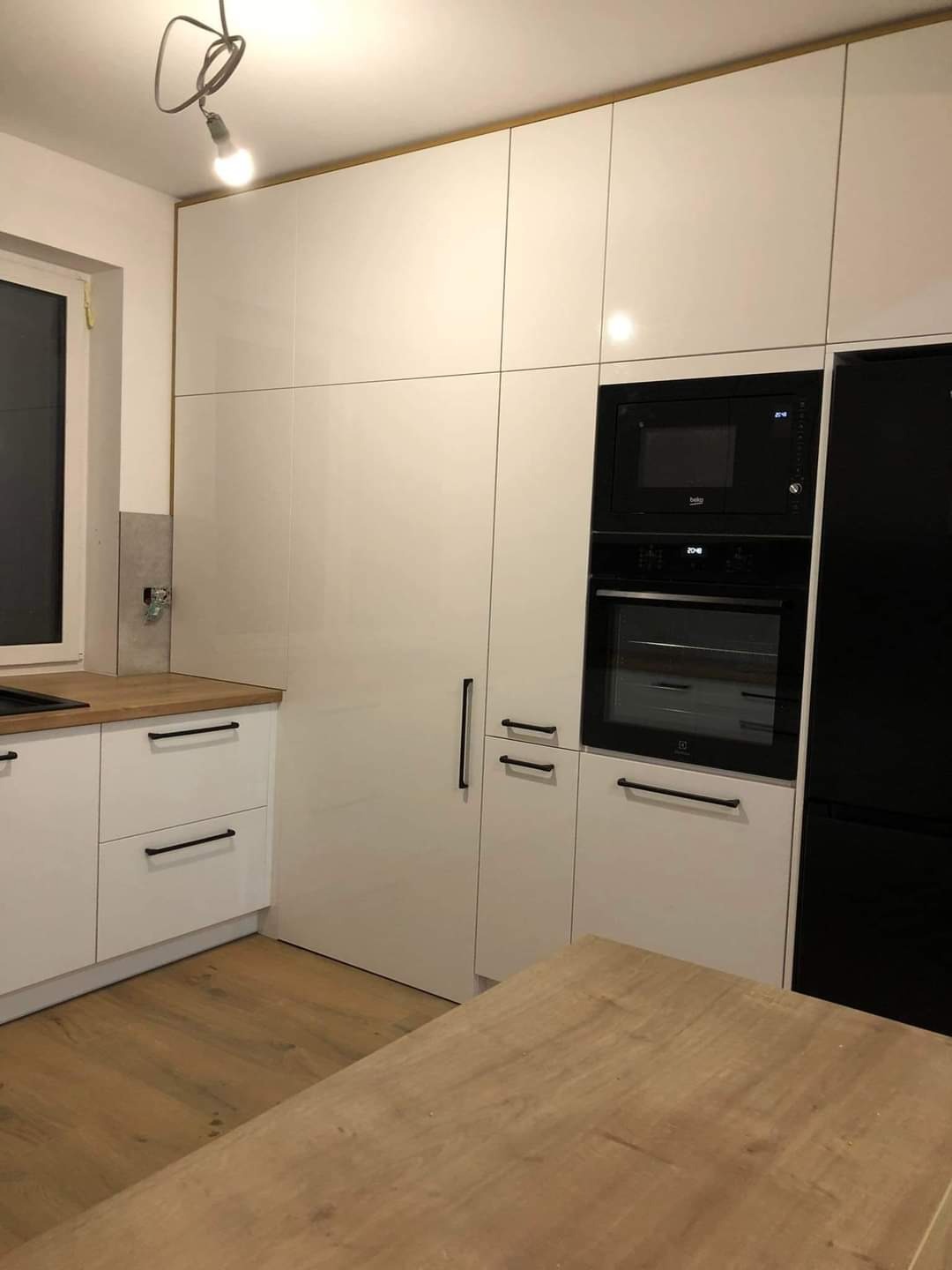 Nowoczesna biała kuchnia na wymiar z czarnymi uchwytami i zabudowanym sprzętem AGD, widoczne elementy wykończeniowe w kolorze drewna.