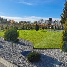 Ujęcie z poziomu gruntu na zadbany trawnik z automatycznym systemem nawadniania w trakcie pracy, otoczony ozdobnymi krzewami i kamieniami, z widocznym domem w tle.