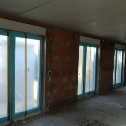 Surowy stan wykończeniowy: rząd okien z niebieskimi ramami, zabezpieczonych folią, z częściowo opuszczonymi, białymi osłonami zewnętrznymi, w murze z cegły dziurawki.