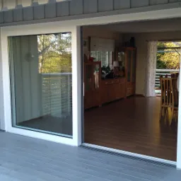 Otwarty, przesuwny system okienny z białymi ramami, widok z zewnątrz na drewniany taras i wnętrze domu z meblami z ciemnego drewna.