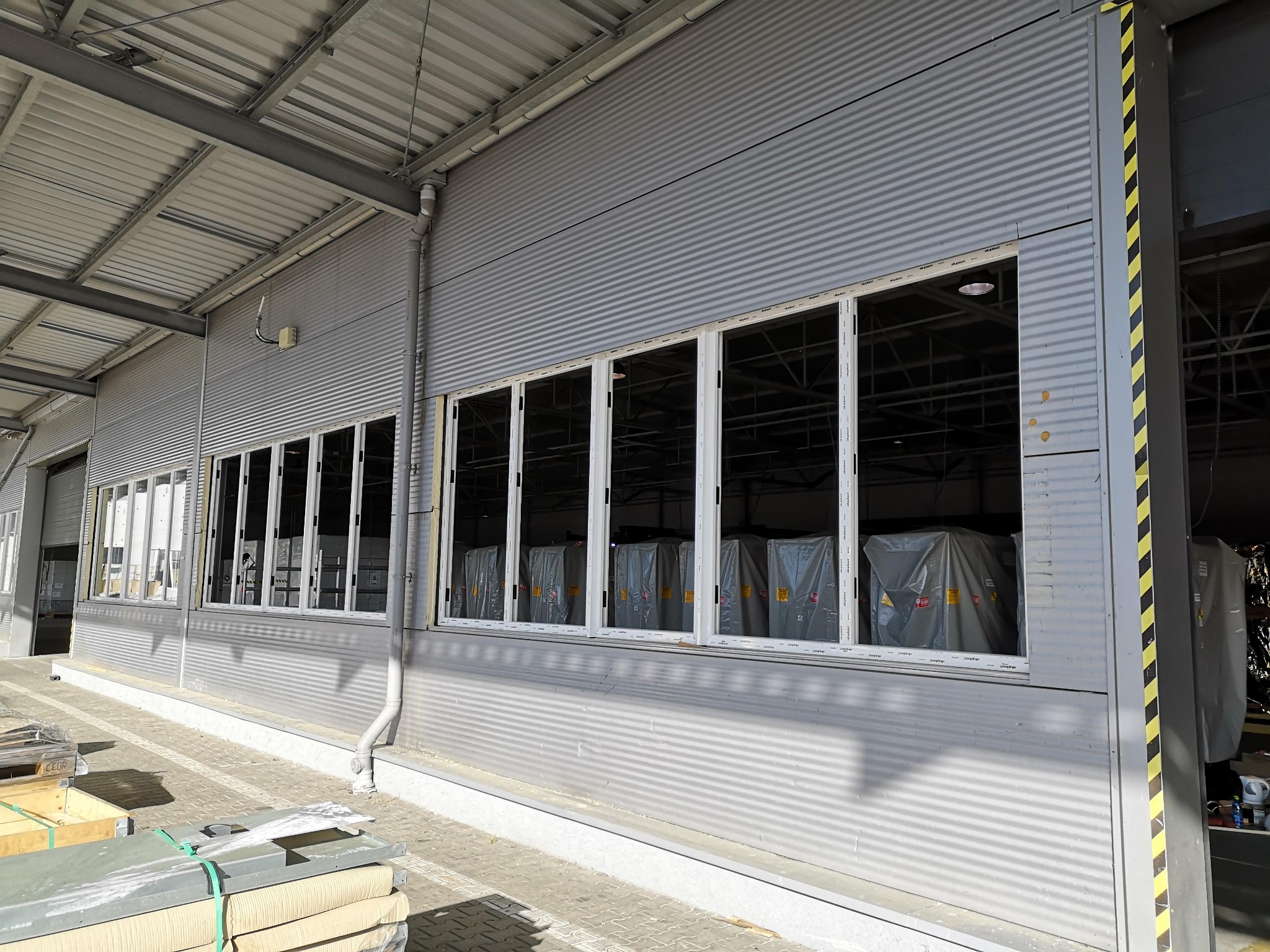 Montaż okien w hali przemysłowej z szarej blachy falistej, widoczne okna z białymi ramami, część z nich zasłonięta folią ochronną, palety z elementami stolarki okiennej na pierwszym planie.