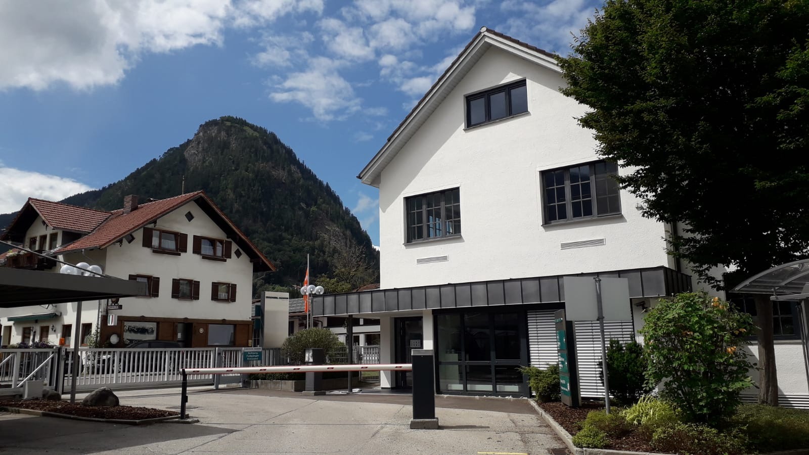 Nowoczesny budynek z białymi ścianami i ciemnymi oknami, widok z zewnątrz, w tle góra porośnięta lasem i budynek z czerwoną dachówką, szlaban przy wjeździe.