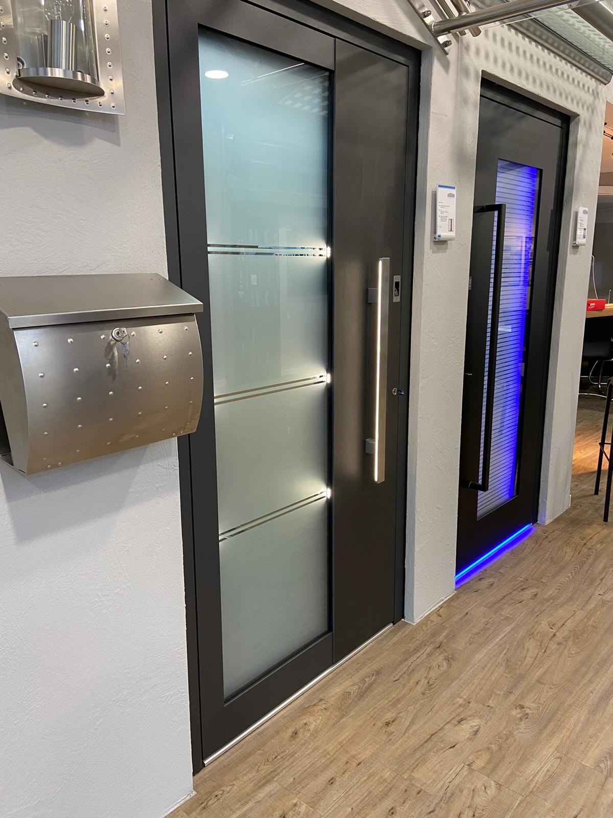 Nowoczesne drzwi wejściowe z matowym szkłem, podświetlanym uchwytem i metalową skrzynką na listy w stylu industrialnym, obok drzwi z niebieskim podświetleniem LED.