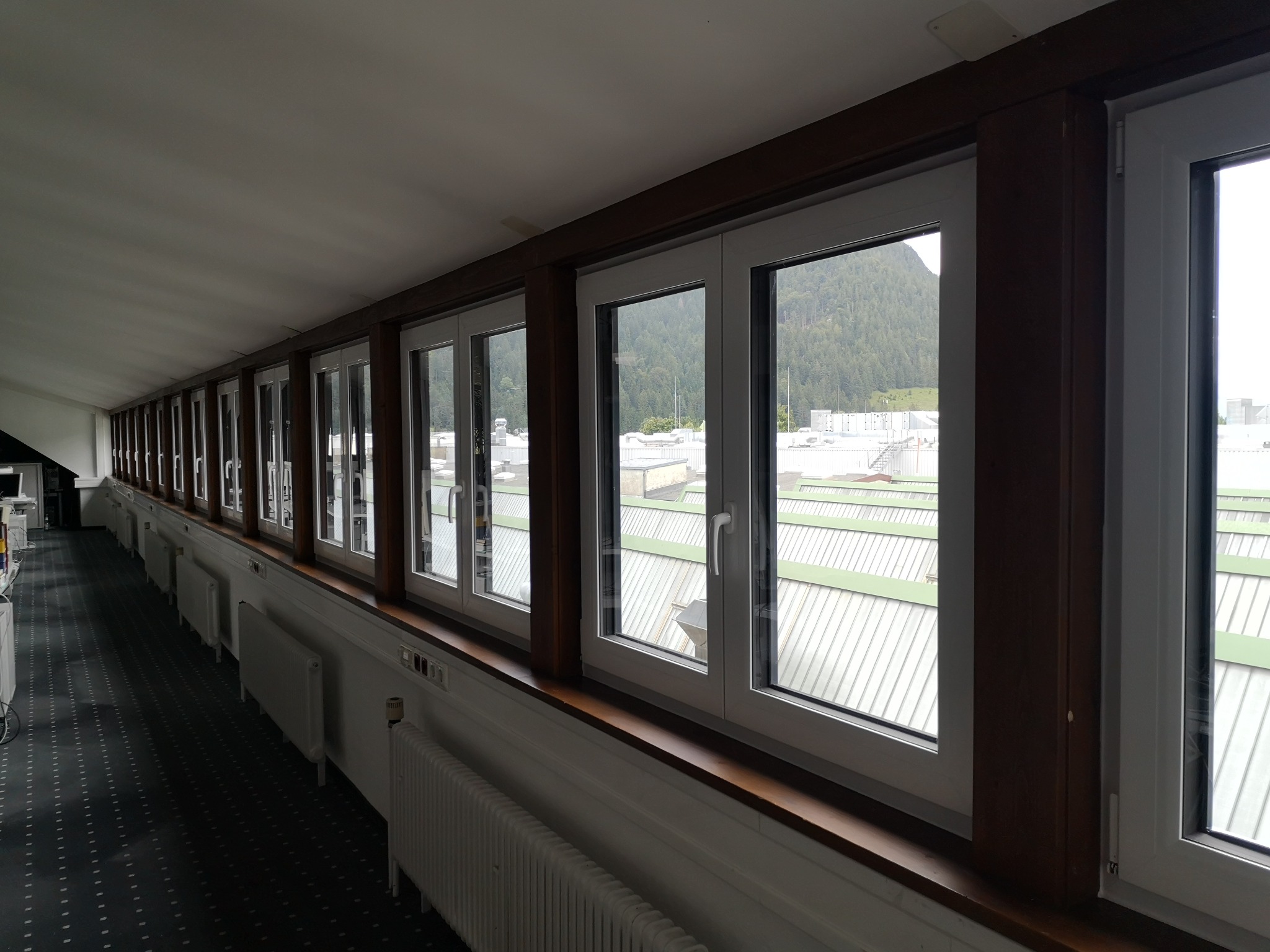 Długi korytarz z rzędem białych okien z widokiem na góry i budynki z blachy falistej, brązowe drewniane ramy okienne, grzejniki pod oknami.